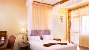 Quarto de hotel com cama, mesa de cabeceira, luminária, cadeira e janela com cortinas.