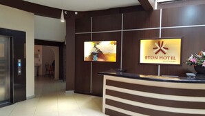 Uma área de recepção de hotel com design moderno, com balcão de atendimento, uma televisão montada e uma placa do Hotel Eton.