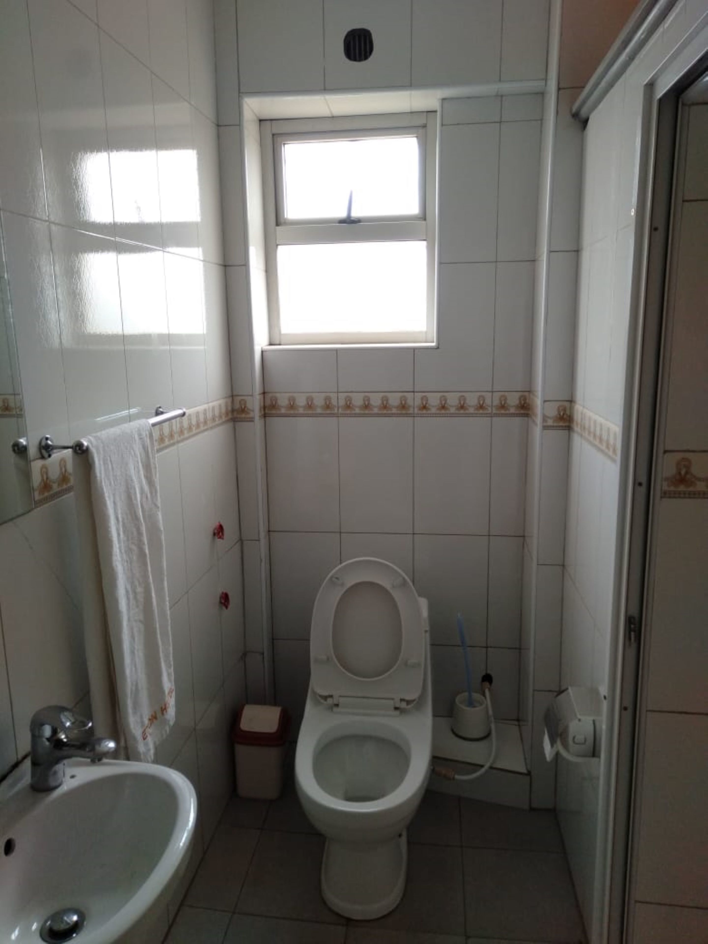 A bathroom with a toilet, sink, and shower. There is a window above the toilet and a towel hanging on the wall.