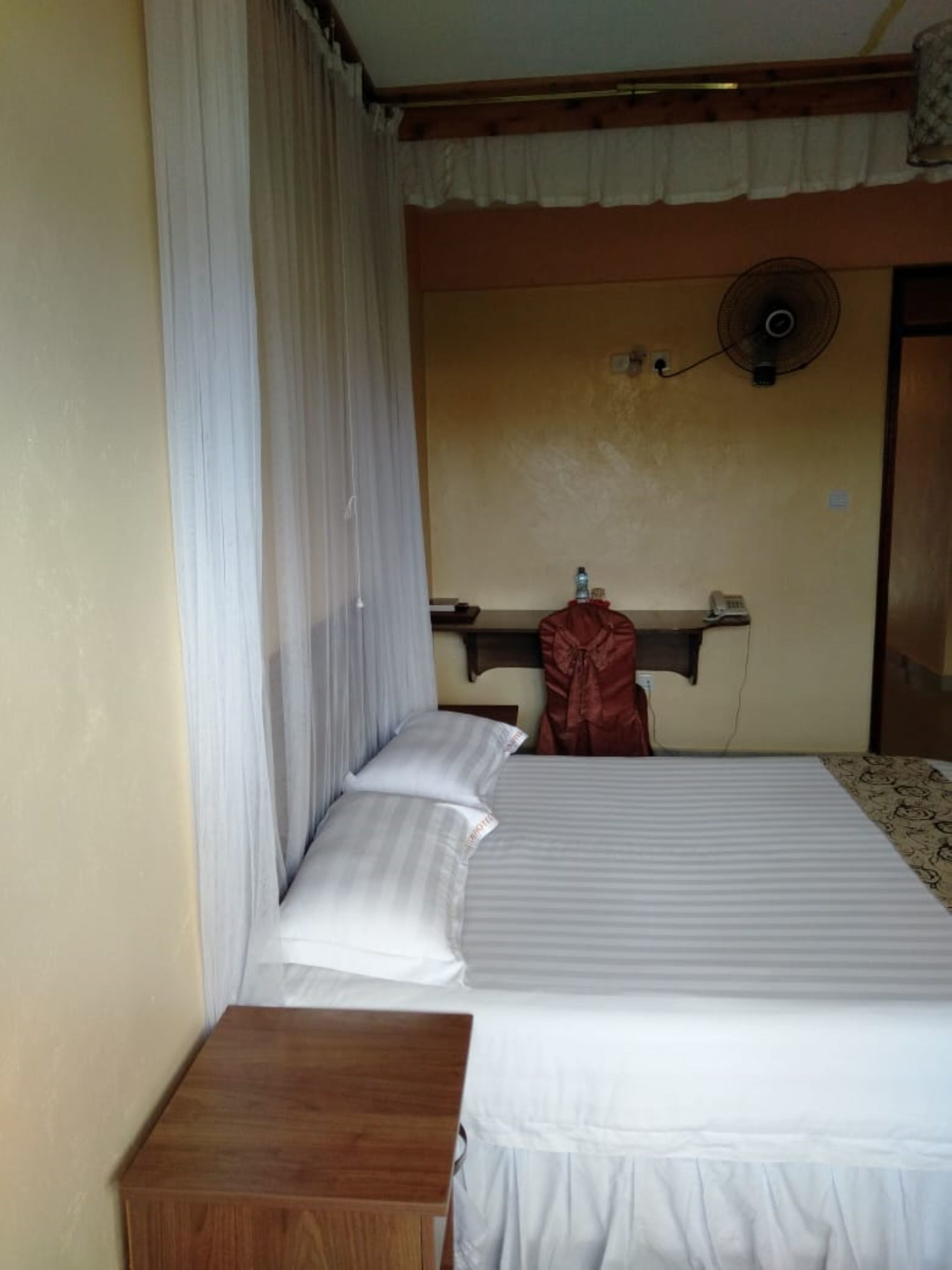 A single bed with white bedding, a wooden nightstand, a fan, and a mirror on the wall.