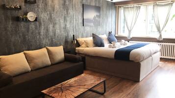 Ein modernes Hotelzimmer mit einer dunkelgrauen, strukturierten Wand, einem großen Bett mit weißer Bettwäsche und blauen Kissen, einem dunkelbraunen Sofa, einem Holztisch und einem großen Fenster mit Vorhängen.