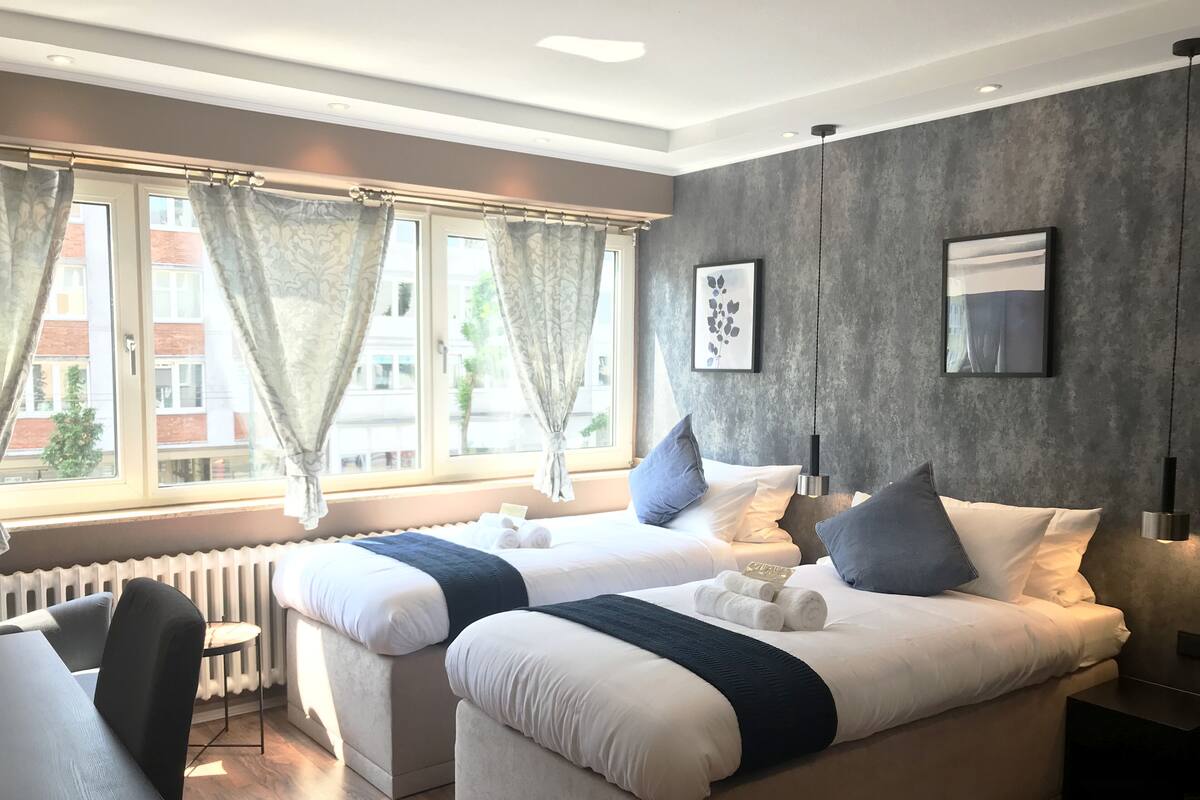 Quarto de hotel moderno com duas camas, uma janela grande com cortinas, um radiador e vista para edifícios.