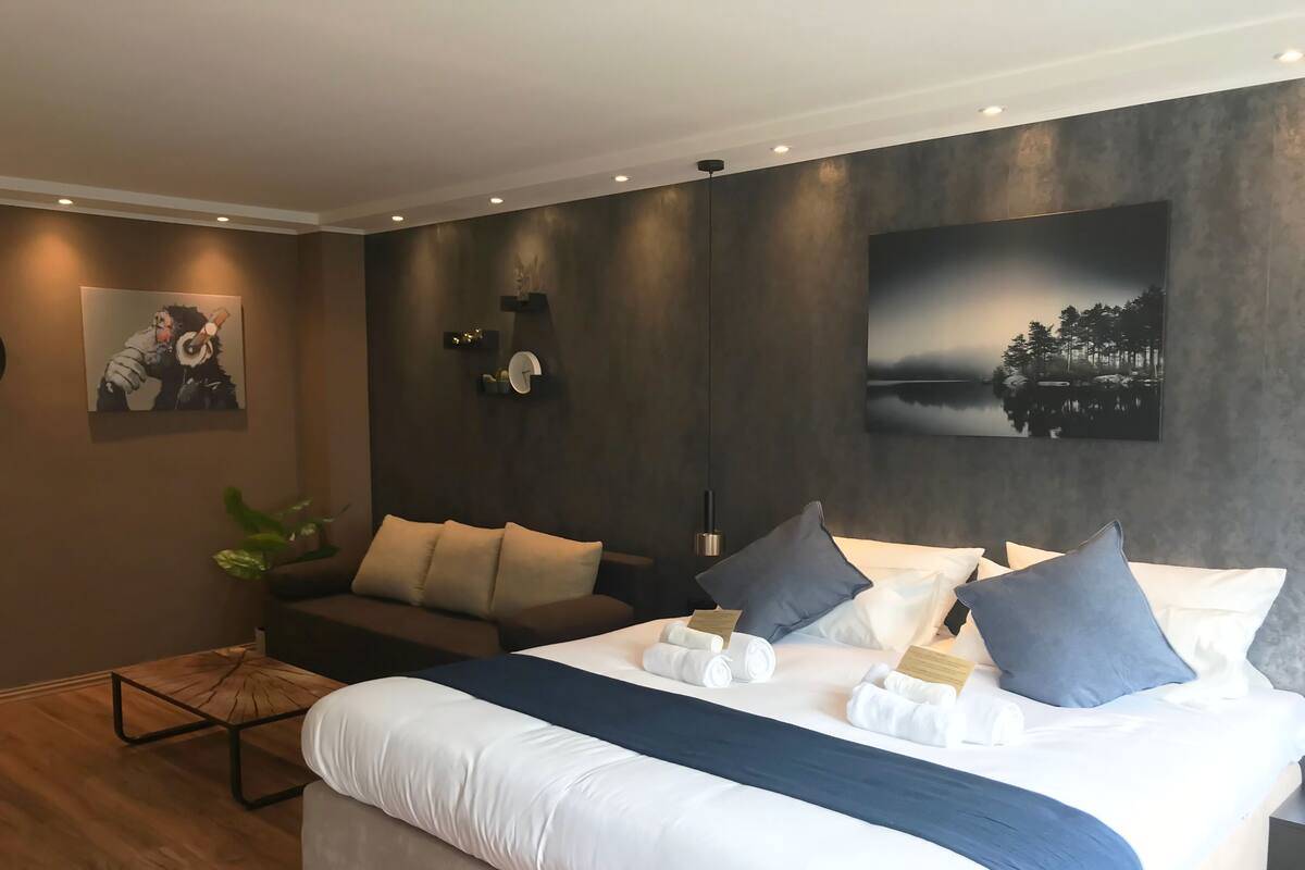 Quarto de hotel moderno com uma cama grande, um sofá e uma pequena mesa. Há quadros nas paredes e um relógio.
