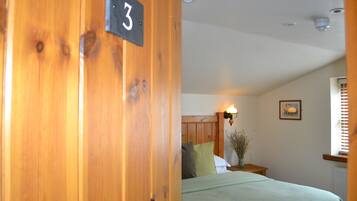 A room with a wooden door marked with the number 3, a bed with a green bedspread, a nightstand with a vase of flowers, and a wall-mounted lamp.