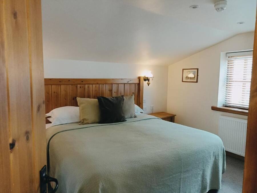 A bedroom with a wooden headboard, a bed with a green blanket, a bedside lamp, and a window with blinds.