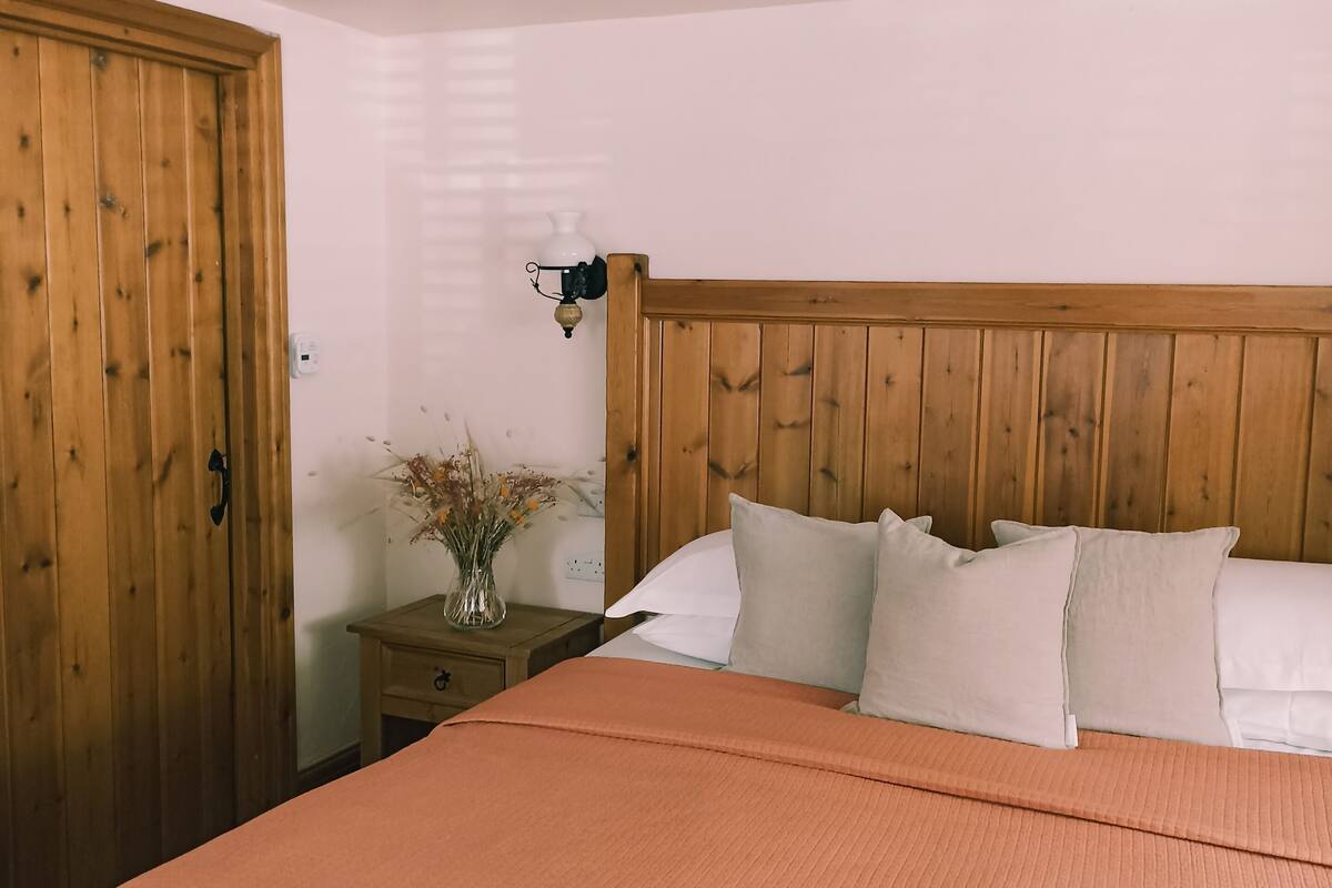 A bedroom with a wooden headboard, a bed with white pillows, a nightstand with a vase of dried flowers, and a wooden wardrobe.