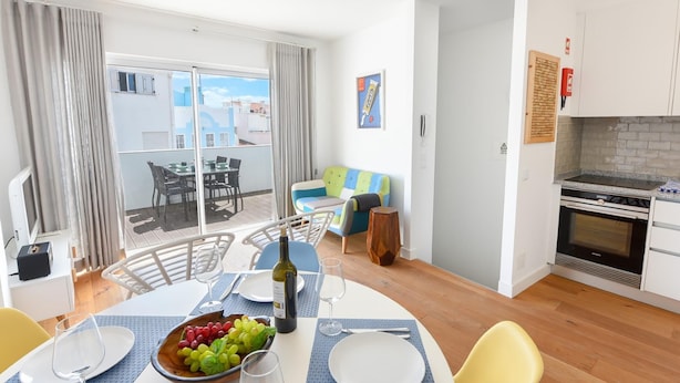 Een eetgedeelte met een tafel gedekt voor twee personen, een fruitschaal en een fles wijn. Er is een balkon met uitzicht op gebouwen en een tafel met stoelen. Achtergrond toont een keuken met een fornuis en kastjes.