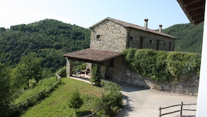 Un bâtiment en pierre, surmonté d’un toit en tuiles, entouré d’une végétation luxuriante et de collines.