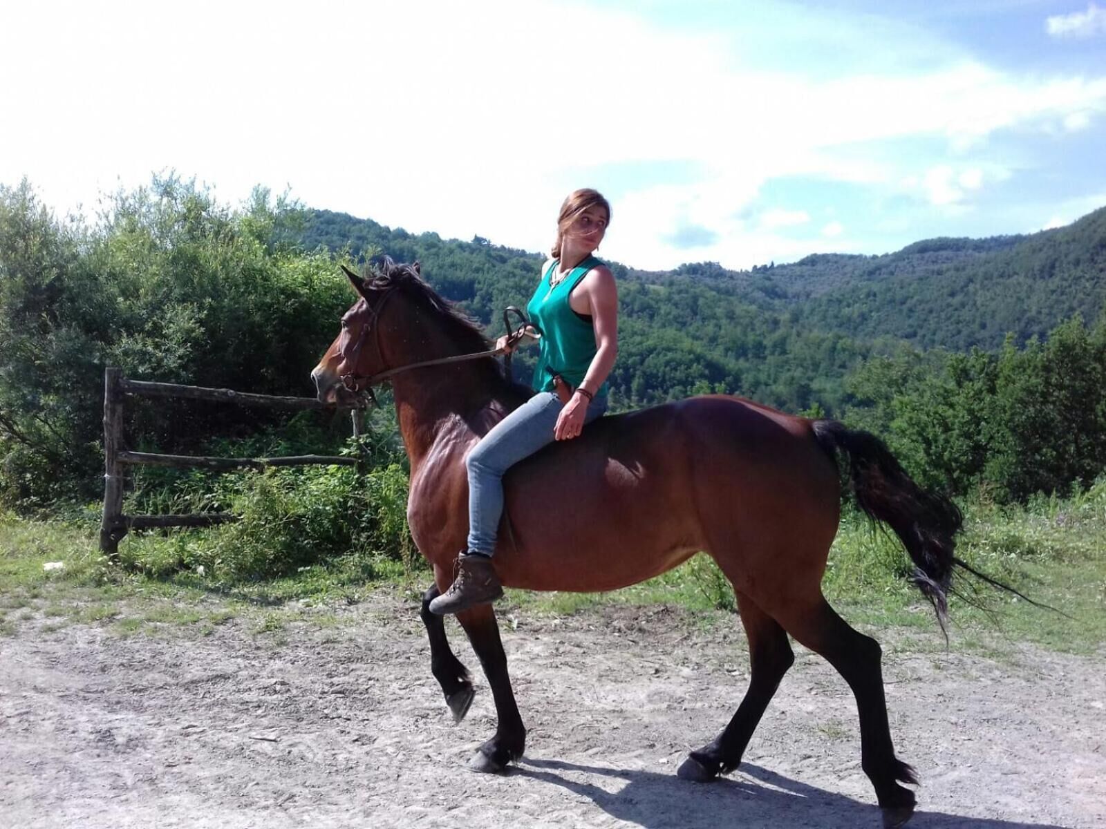 Una persona montando a caballo por un sendero de tierra con una cerca de madera y vegetación al fondo.