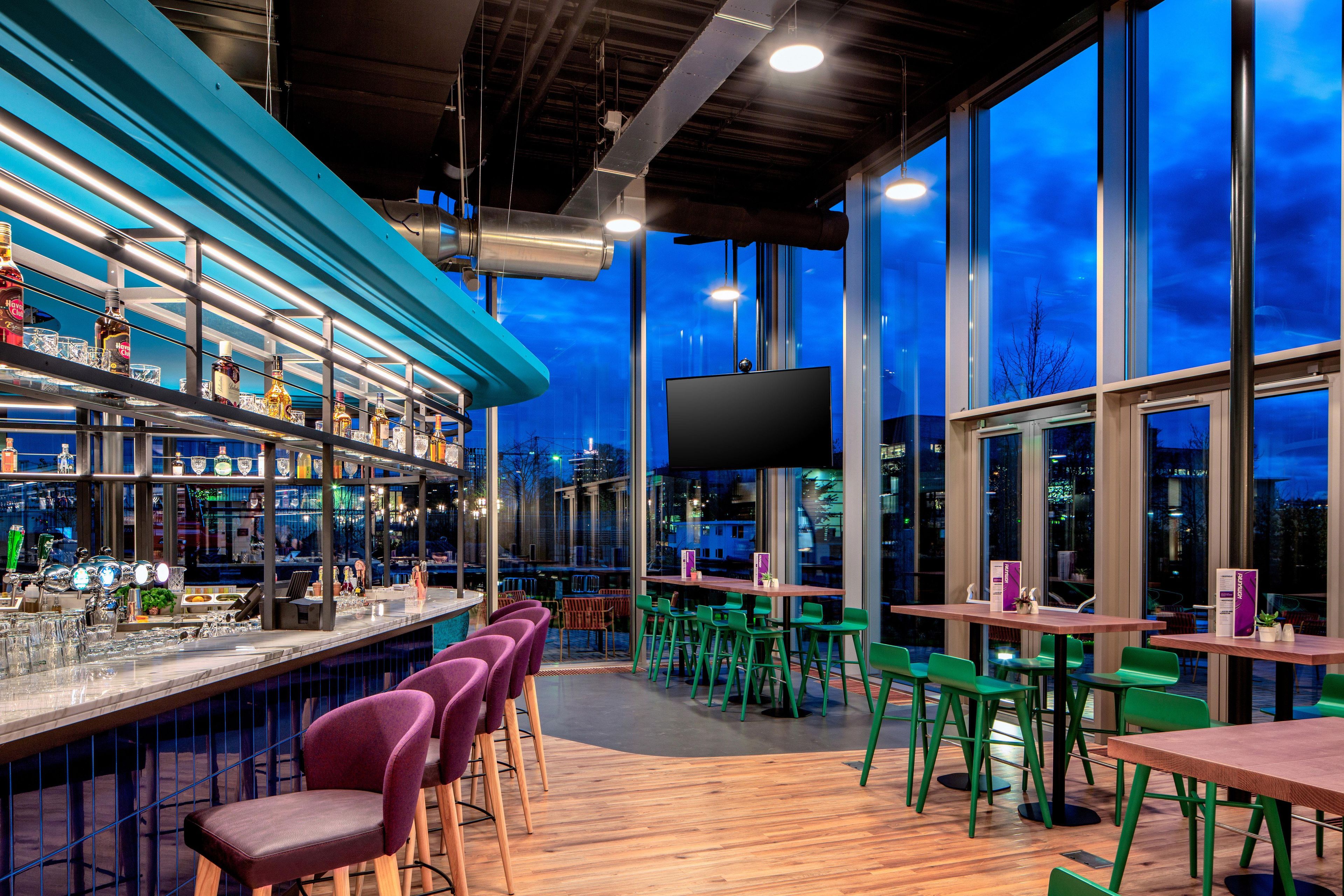 A bar area with a long counter, multiple stools, and a large window offering a view of the cityscape at night.