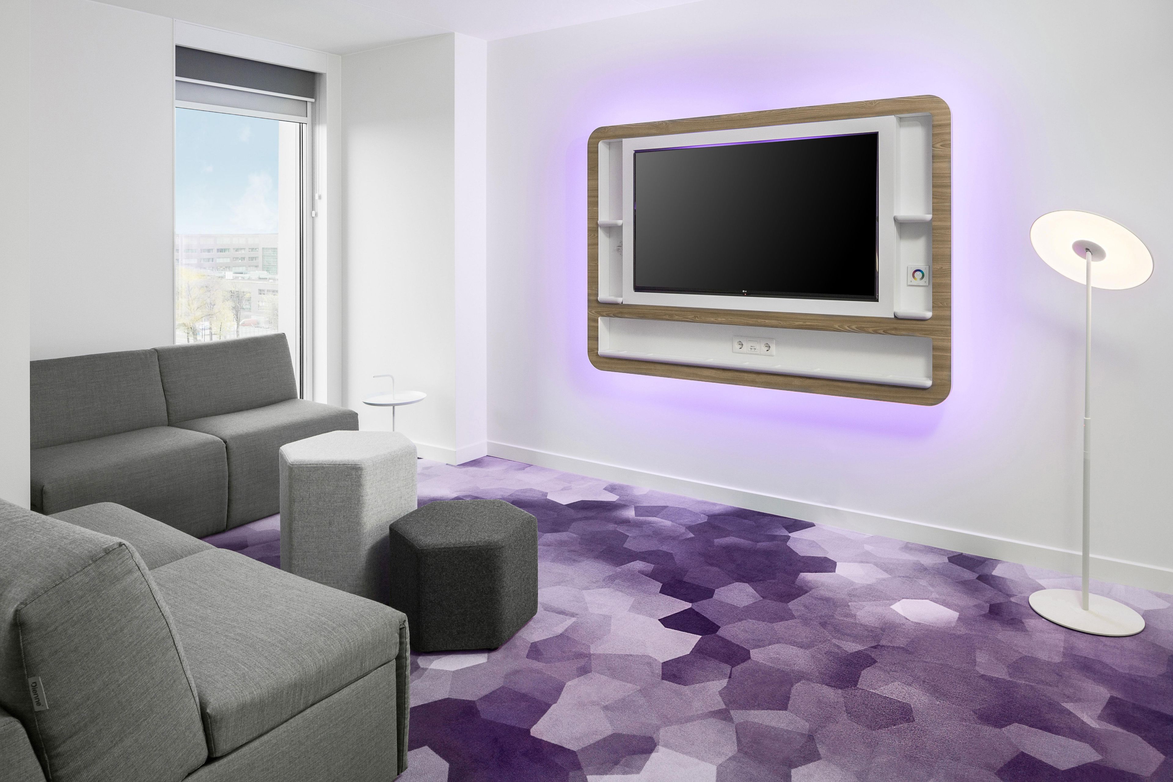 Una sala de estar moderna con un televisor de pantalla plana montado en la pared, sofás grises, un otoman redondos y una alfombra púrpura con motivos geométricos.