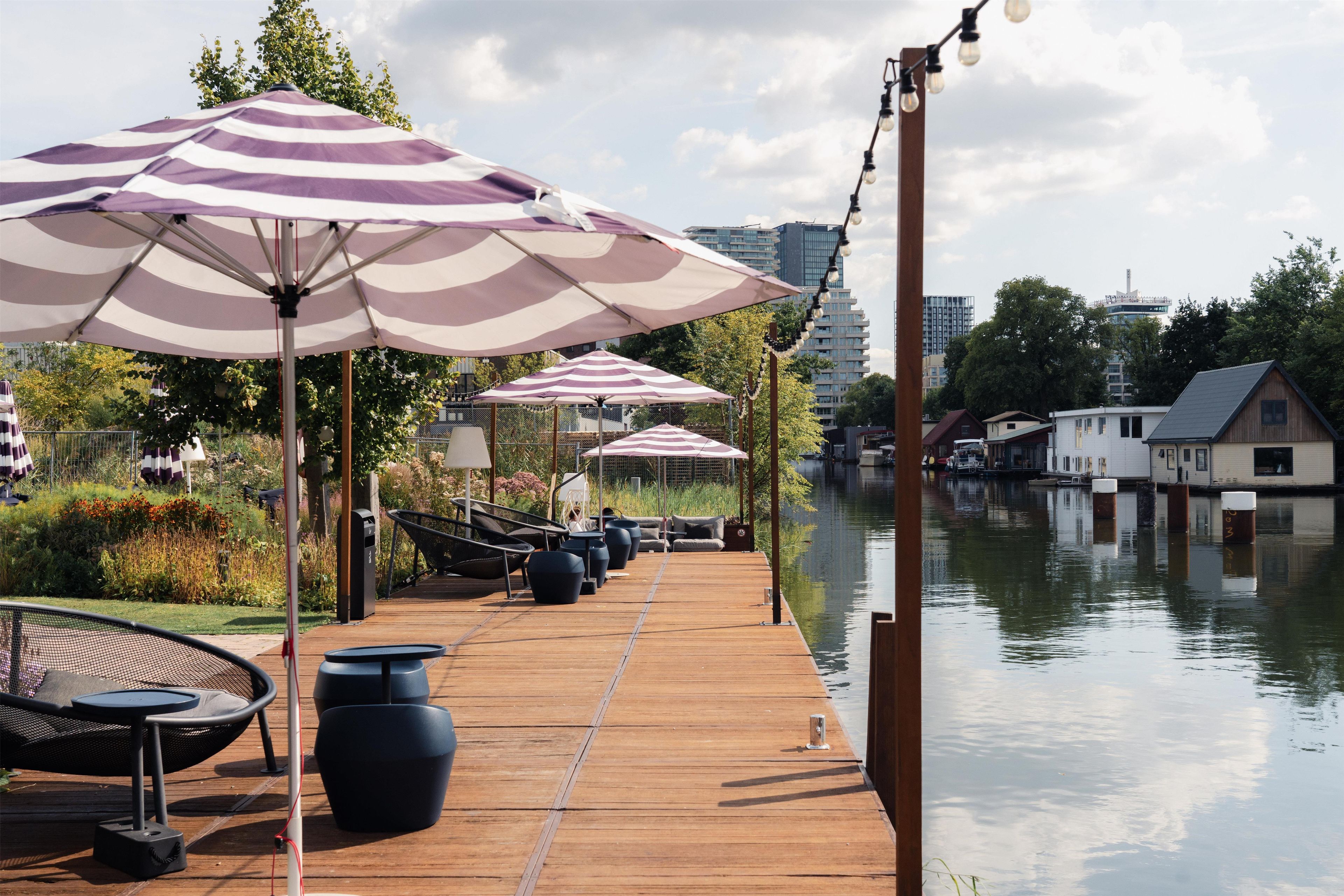 Une terrasse en bois avec des sièges et des parasols, donnant sur une étendue d’eau calme, avec des bâtiments en arrière-plan.