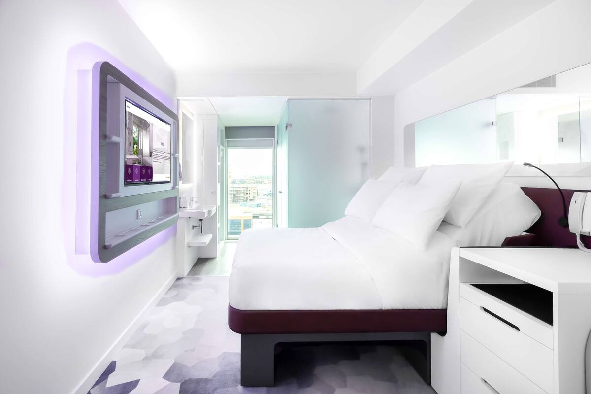 Une chambre d’hôtel moderne avec un grand lit, une table de chevet, un téléviseur intégré et une vue sur la ville par la fenêtre.