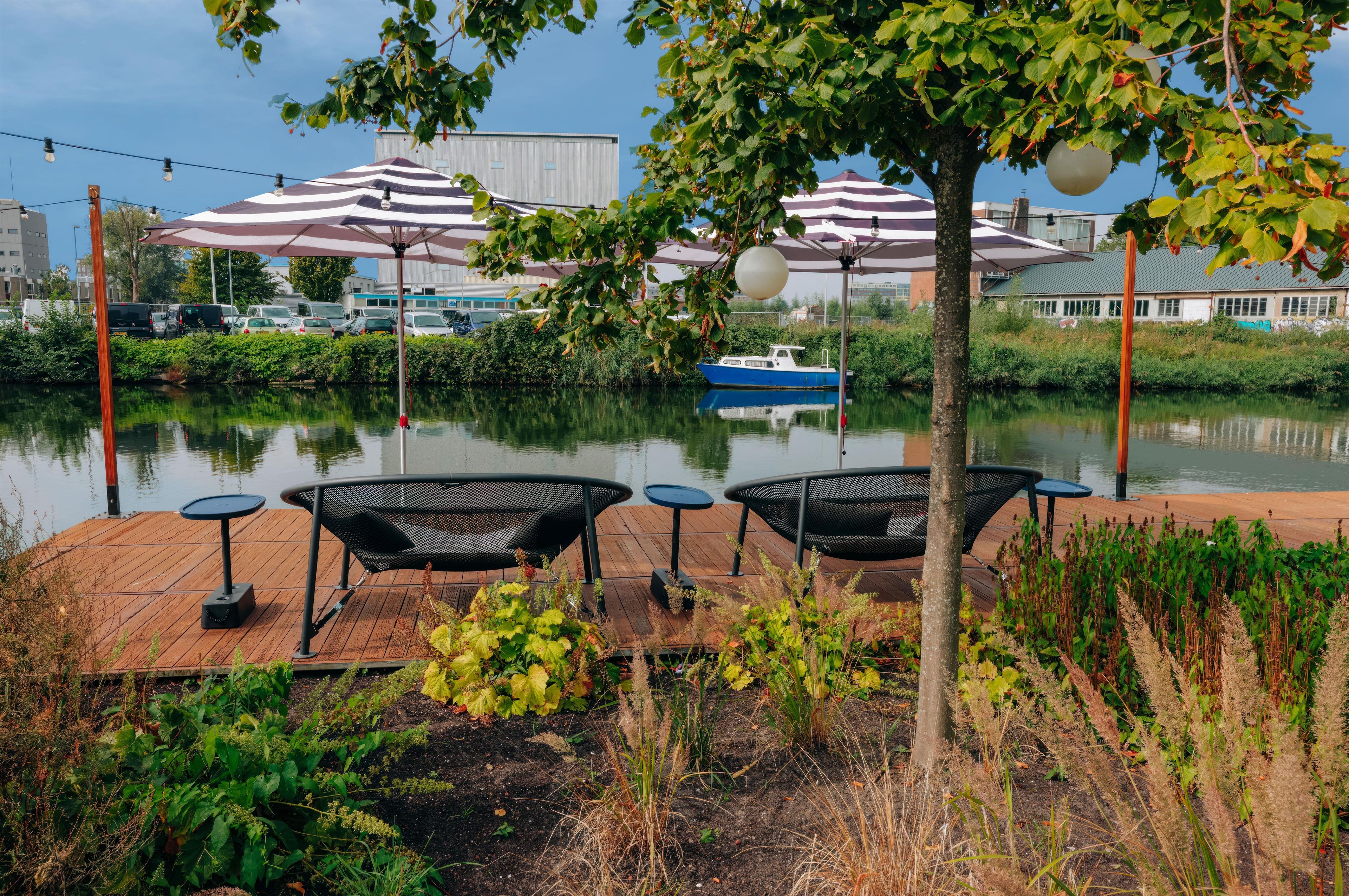A riverside deck with outdoor seating and umbrellas, a boat on the water, and industrial buildings in the background.