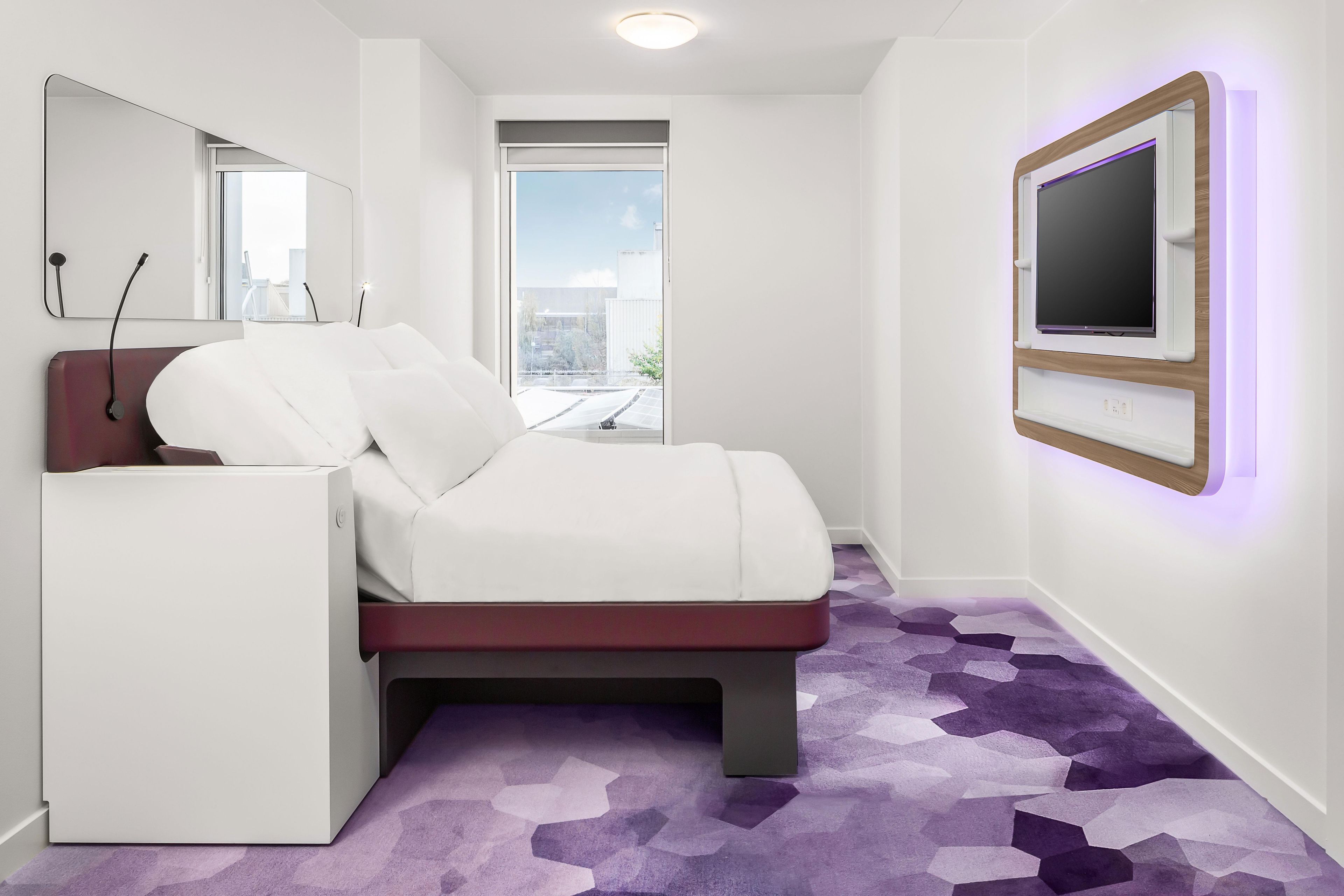 A modern bedroom with a bed, a small white cabinet, a large mirror, and a television mounted on the wall.