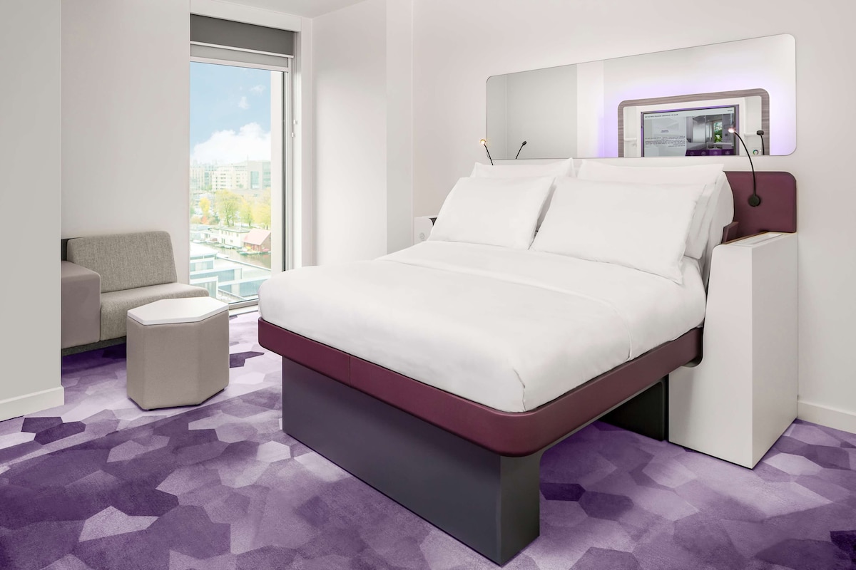 Una habitación de hotel moderna con una cama grande, un pequeño sofá, una mesita de noche y un amplio ventanal con vistas a la ciudad.