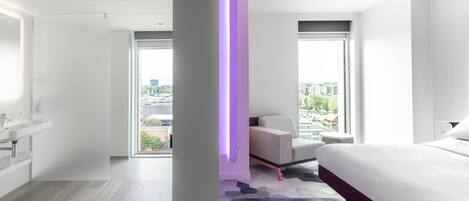 A modern hotel room with a large bed, a desk, a chair, and a view of the city.