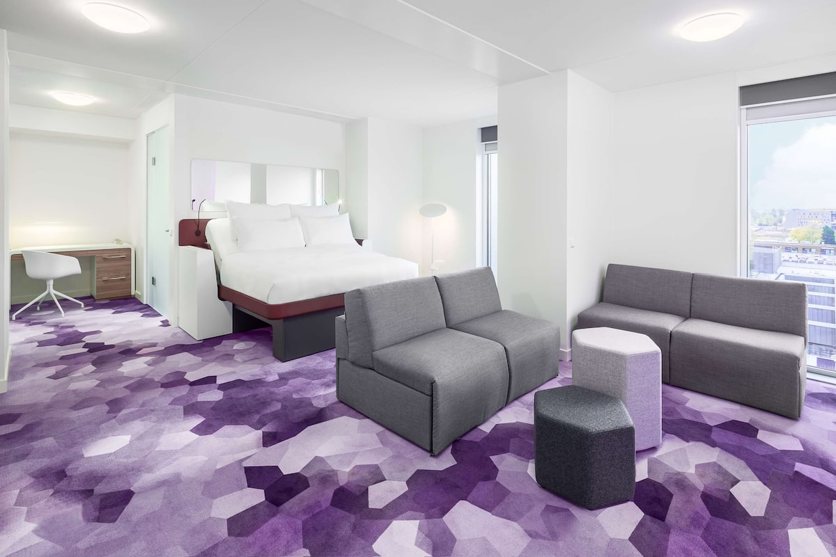 Une chambre d’hôtel moderne avec un lit, un canapé gris, un pouf et un bureau avec une chaise.