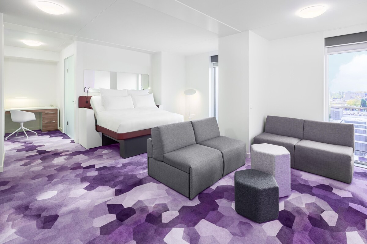 Una habitación de hotel moderna con cama, dos sofás, un escritorio y un ventanal con vistas a la ciudad.