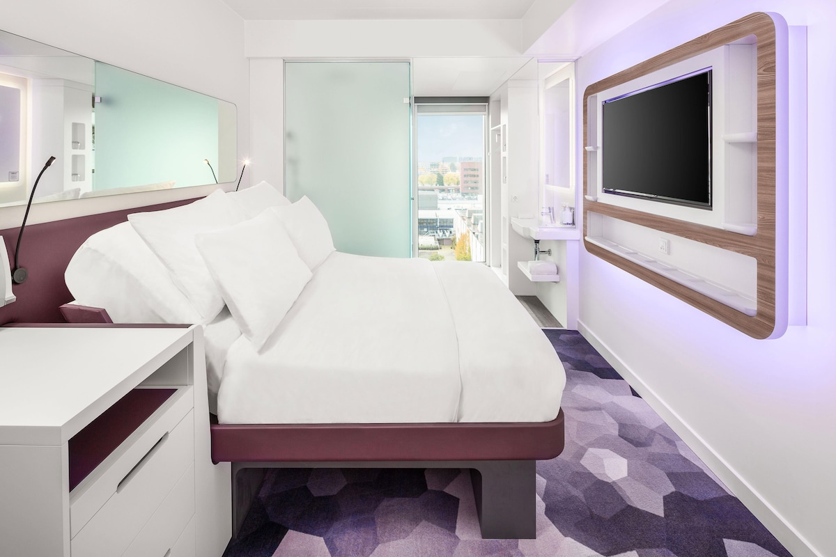 Habitación de hotel moderna con cama, escritorio, televisor y baño visible a través de un vidrio.