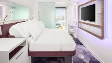 Habitación de hotel moderna con cama, escritorio, televisor y baño visible a través de un vidrio.