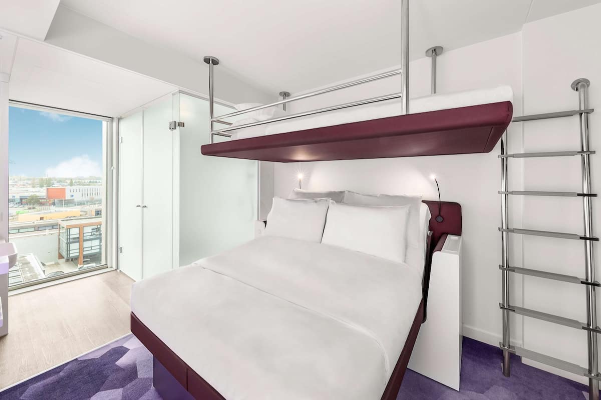 Une chambre d’hôtel moderne avec un grand lit, une échelle intégrée et une vue sur la ville grâce à une grande fenêtre.