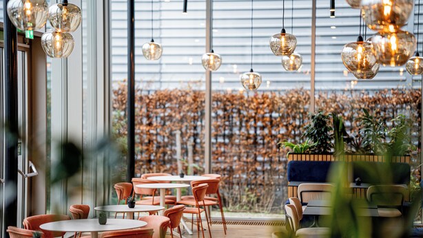Un comedor moderno con mesas redondas y sillas naranja, ventanas amplias y lámparas esféricas colgantes.