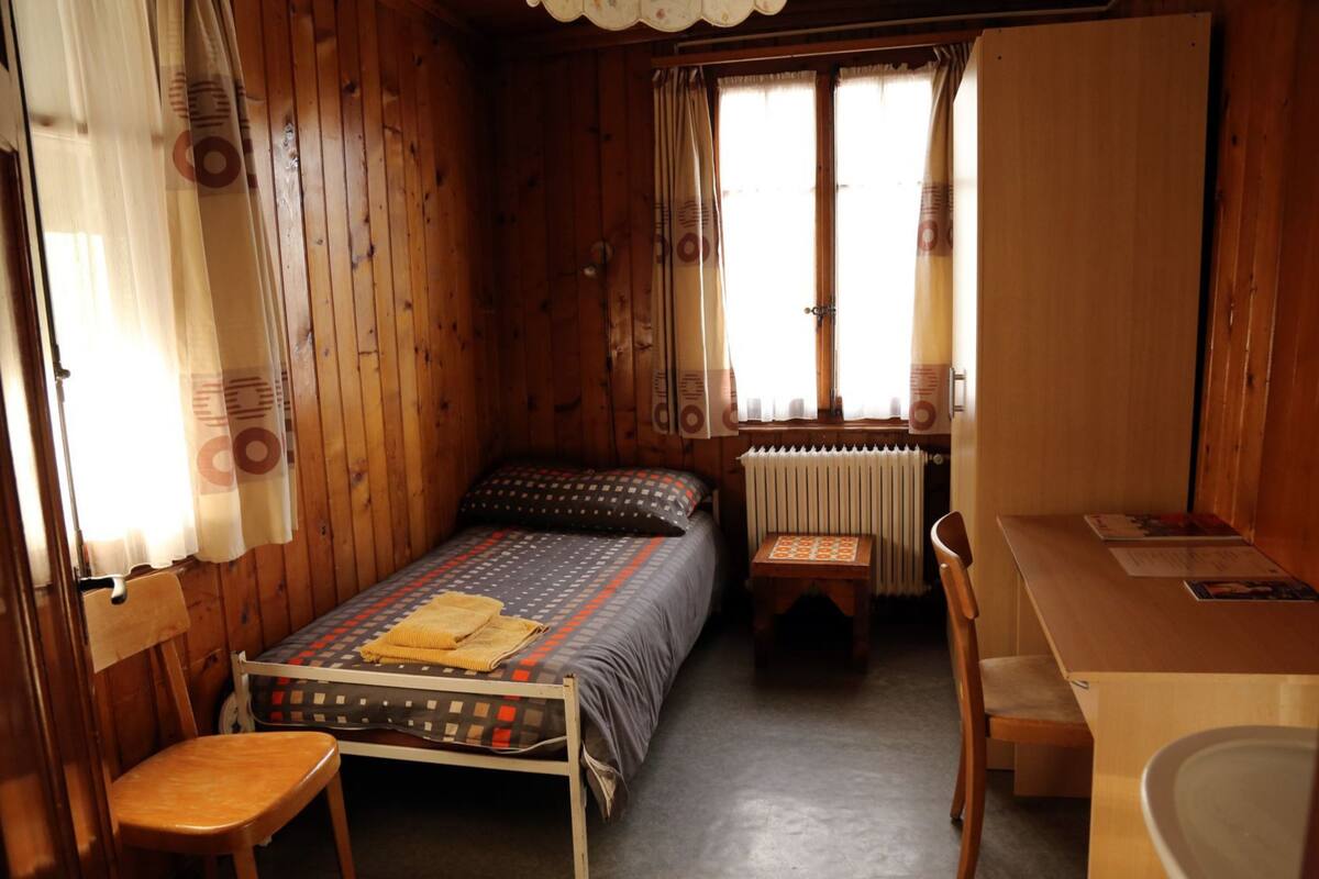 Una habitación pequeña y acogedora con paredes de madera y una cama individual con colcha estampada. Hay una mesita y una silla, y un radiador debajo de una ventana.