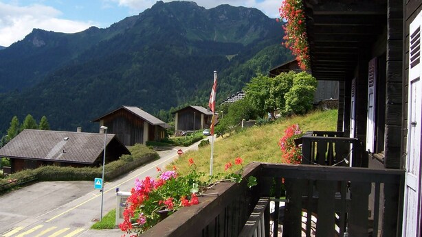 Un pueblo de montaña con casas de madera tradicionales, un asta de bandera y vistas despejadas de las montañas circundantes.