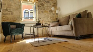 A cozy living room with a stone wall, a window with curtains, a green armchair, a beige sofa, and a small table with a plant. - The Suites at Central Split (Split)