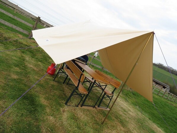 Tienda de campaña con toldo color beige y estructura negra, montada en un campo cubierto de césped con mesas de picnic y un cilindro de gas rojo cerca.