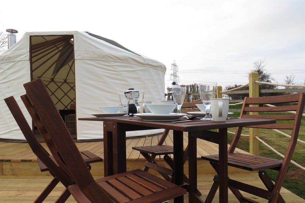 A wooden deck with a table set for dining, surrounded by wooden chairs. A white tent is visible in the background.