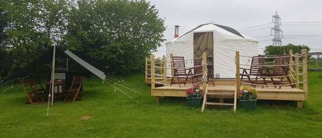 A white tent with a wooden deck and seating area, surrounded by greenery.