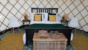 Cama com cabeceira preta, almofadas brancas e mostarda, e manta verde. Duas mesas de cabeceira com luminárias e pequenas plantas em vasos. Um cesto de palha e uma caixa estão sobre a cama.