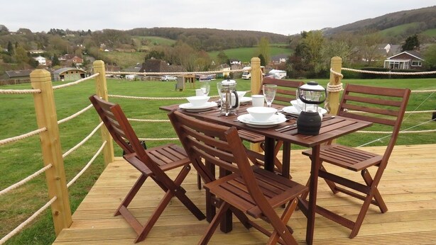 Um deck de madeira com uma mesa posta para refeições, rodeado por cadeiras, com vista para uma paisagem rural com casas e colinas.