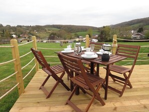 Una terraza de madera con una mesa dispuesta para comer, rodeada de sillas, con vistas a un paisaje rural con casas y colinas.