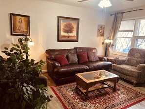 Una sala de estar con un sofá de cuero marrón, un sillón estampado, una mesa de centro de madera y un tapete con motivos rojos y dorados. Hay cuadros colgados en la pared y una planta en la esquina.