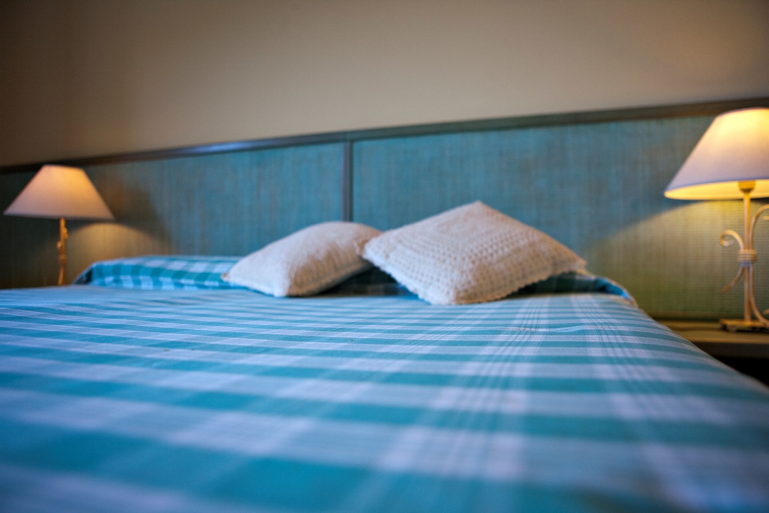 Ein Bett mit einer blau-weißen karierten Tagesdecke und zwei weißen Kissen. Auf beiden Seiten des Bettes stehen Nachttische mit Lampen und Schirm.