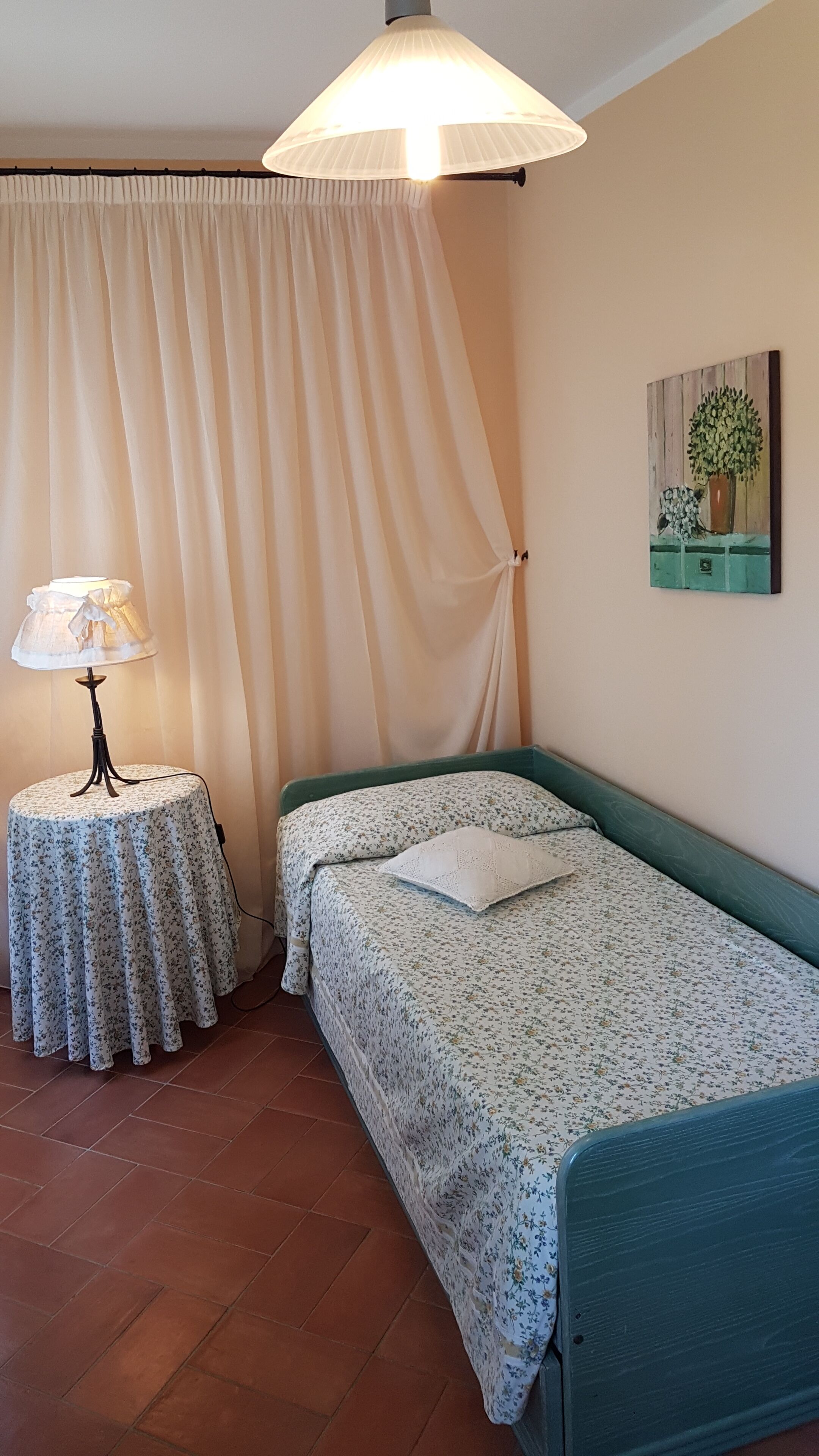 A bedroom with a single bed, a bedside table with a lamp, a curtain, and a painting on the wall.