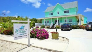 A turquoise two-story house with a porch, surrounded by a sandy area and colorful flowers.