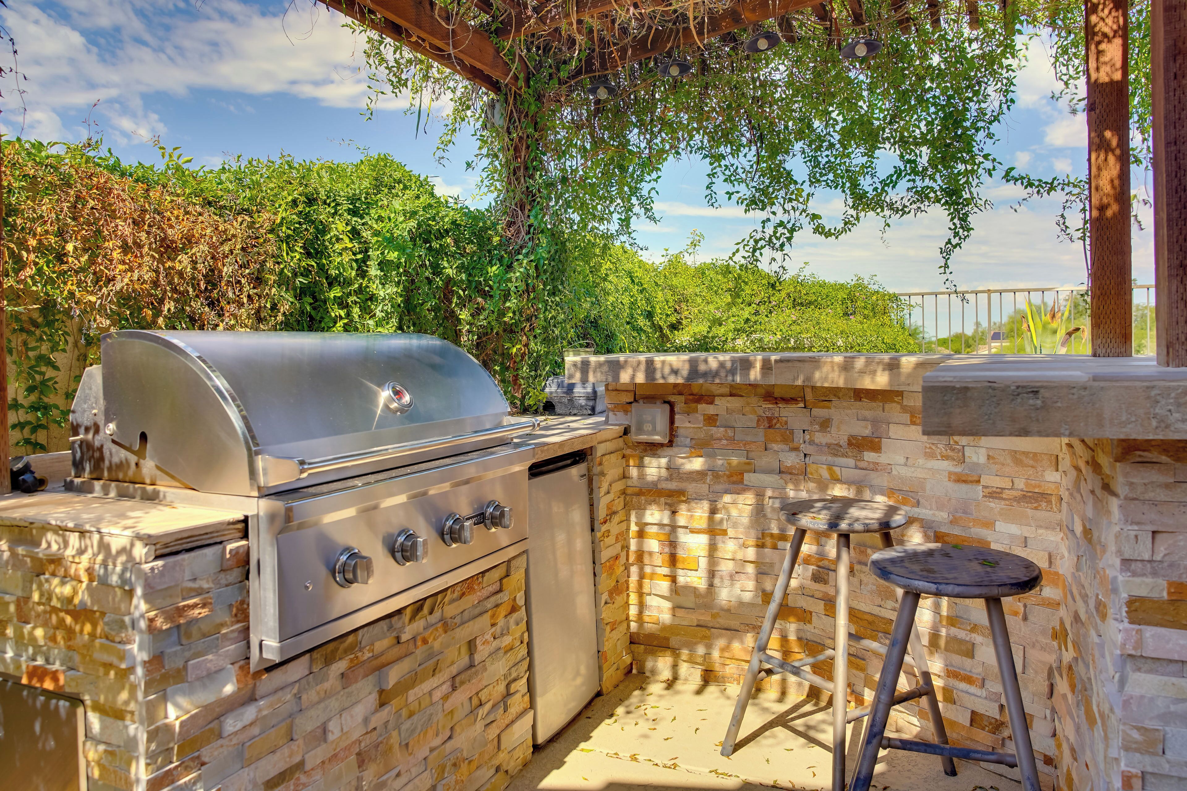 An outdoor kitchen with a built-in grill, a compact refrigerator, and two stools.