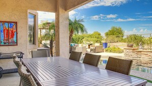 Eine überdachte Terrasse mit Esstisch und Korbstühlen, mit Blick auf einen Swimmingpool und üppige Vegetation.