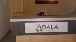 Un comptoir en bois sur lequel est posée une enseigne indiquant « Adala Motel », une peinture représentant un arbre se détachant sur un coucher de soleil, et une petite figurine d’éléphant.