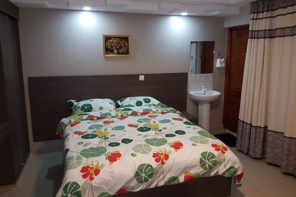 A bedroom with a bed, a bedside table, a wall-mounted mirror, a sink, and a door with curtains.