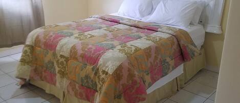 Una cama individual con colcha estampada y almohadas blancas en una habitación con paredes lisas y suelo de baldosas.