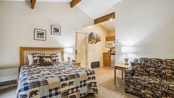 A bedroom with a bed, a sofa, a desk, a chair, a hanging light, a door, and a kitchen area.