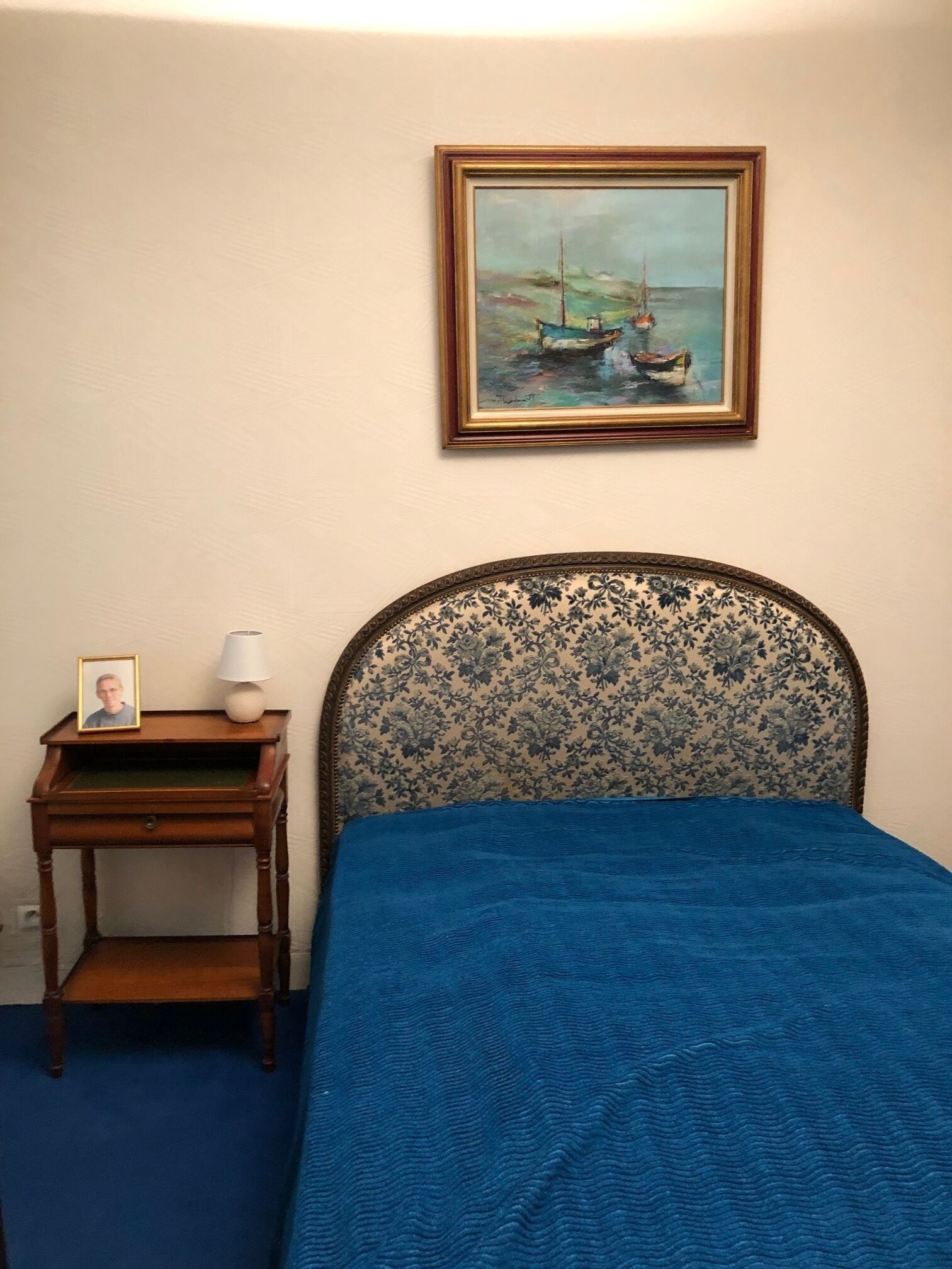 A bedroom with a bed, a nightstand, a framed painting, and a lamp.