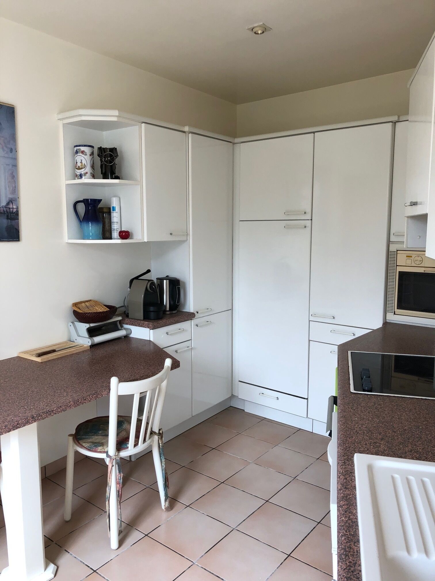 A compact kitchen with white cabinets, a countertop, and a microwave. There is a chair and a small table with a basket and a kettle.
