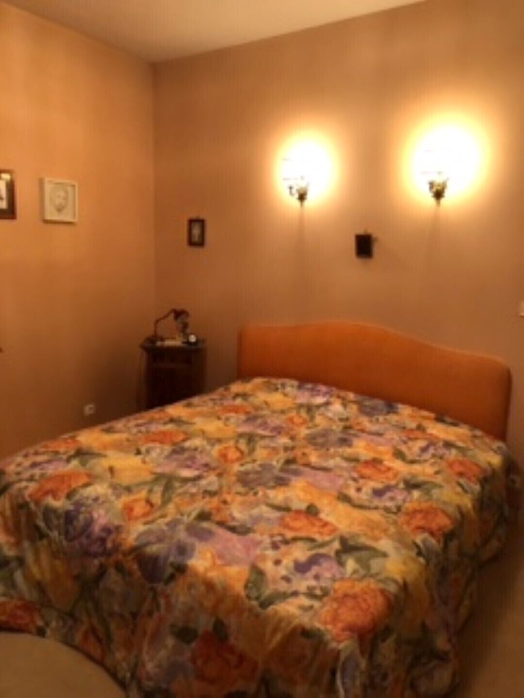 A bedroom with a floral bedspread, two wall-mounted lamps, and a nightstand with a lamp.