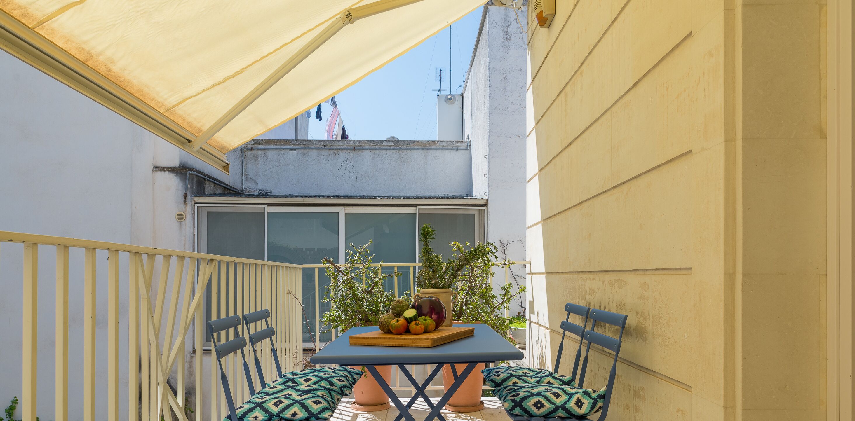 Un pequeño patio al aire libre con una mesa lista para comer, rodeado de sillas y una sombrilla grande que da sombra.